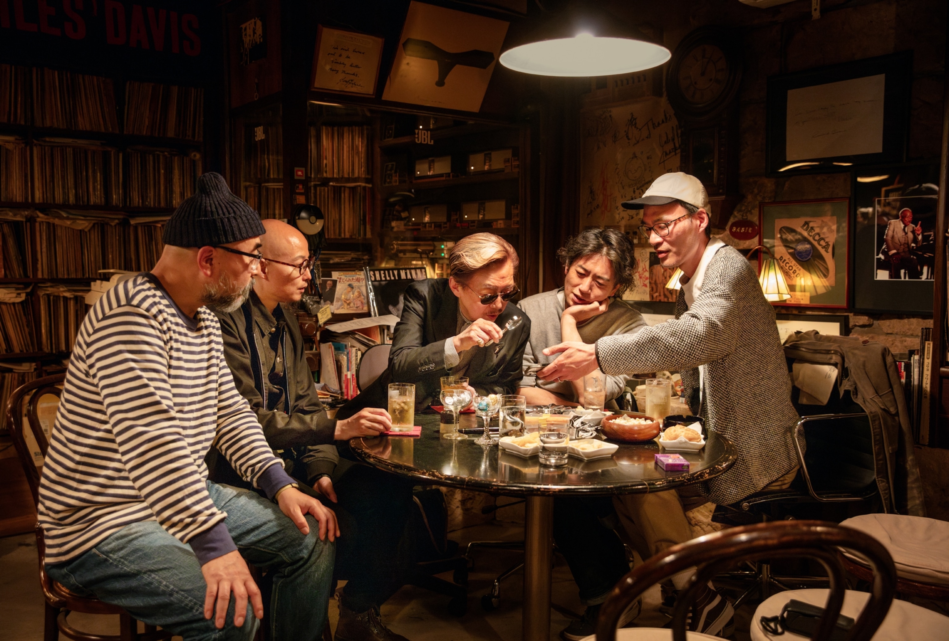 Group of people around table loaded with drinks and snacks looking at something through the magnify glass.